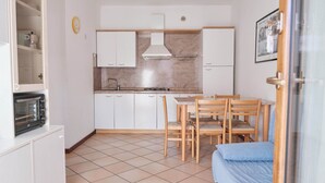 Een kleine keuken met witte kastjes, een gasfornuis en een magnetron. Er is een eettafel met stoelen en een blauwe bank in de hoek.