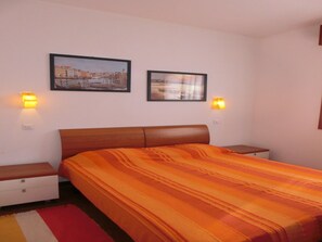 Un dormitorio con una cama, dos mesitas de noche, una lámpara de noche, una alfombra roja y blanca a rayas, y dos luces fijadas a la pared.