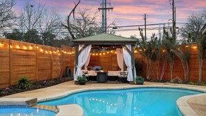 Un patio trasero con piscina, terraza de madera, un cenador con luces colgantes y una chimenea.