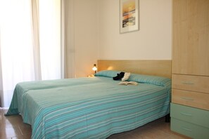 A room with two beds, a nightstand with a lamp, a wooden wardrobe, and a painting on the wall. - Flat with a lovely poolside garden with bbq (Bibione)