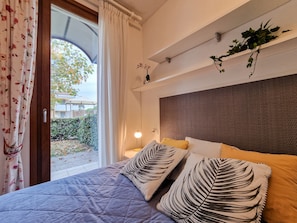 Quarto com cama, almofadas, mesa de cabeceira, abajur, janela com cortinas e planta na parede.