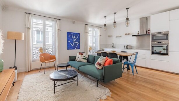 Un apartamento moderno con sala de estar, comedor y cocina. Incluye un sofá, una mesa de comedor y sillas. La cocina tiene hornos empotrados y un ventanal.