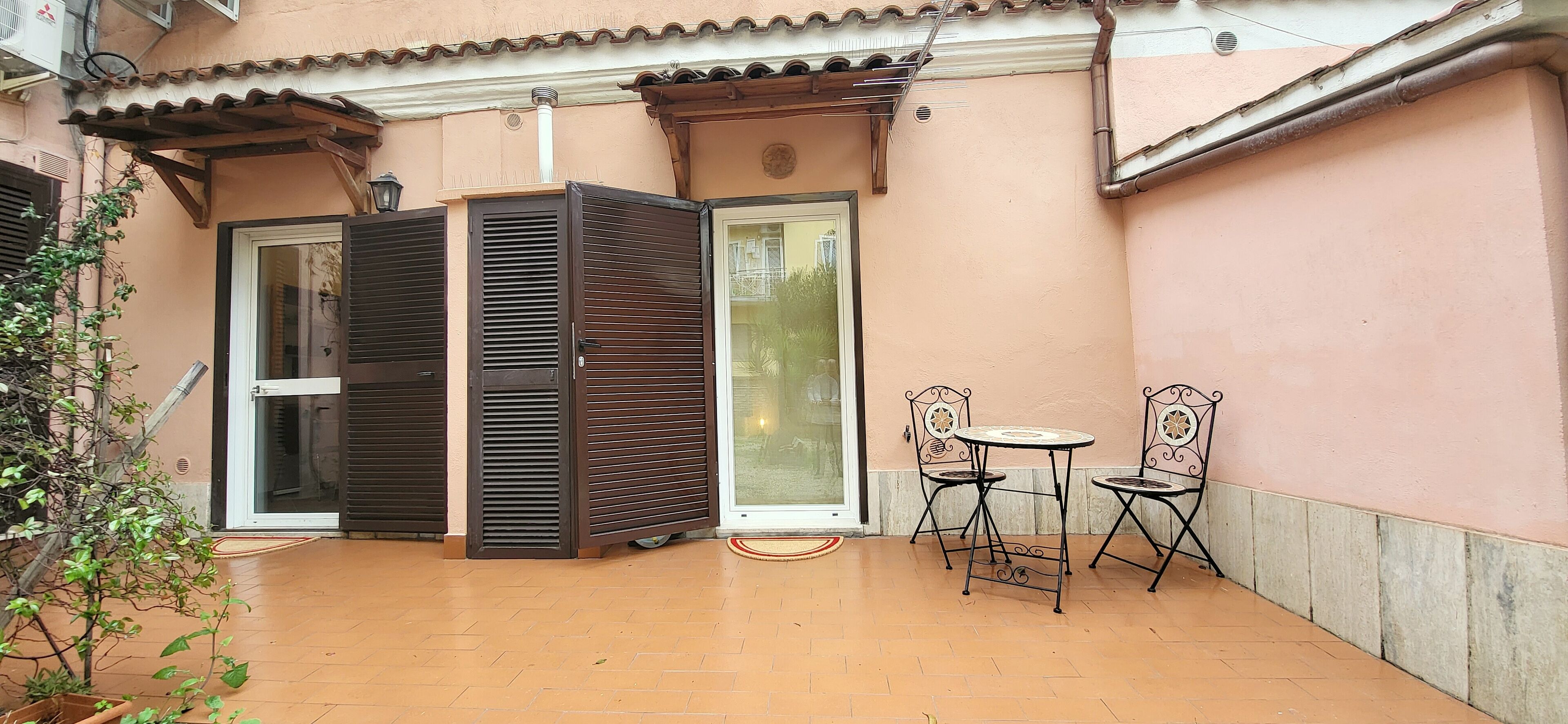 Un patio con pavimento piastrellato, due sedie in metallo, un piccolo tavolino rotondo e una porta scorrevole con finestra.