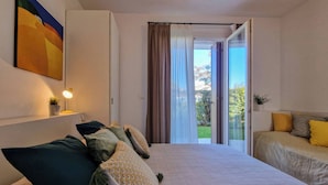 Ein Schlafzimmer mit Bett, Nachttisch mit Lampe, einem Fenster mit Vorhängen und Blick ins Freie.