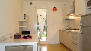 A compact kitchen with a dining area, a refrigerator, and a window with curtains. - Modern one bedroom apartment with solarium (Bibione)