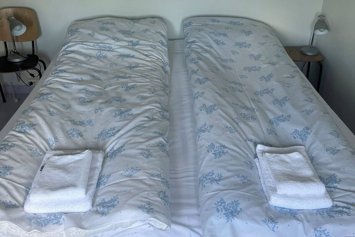 A double bed with floral bedding and two towels, a bedside table with a lamp, and a chair.