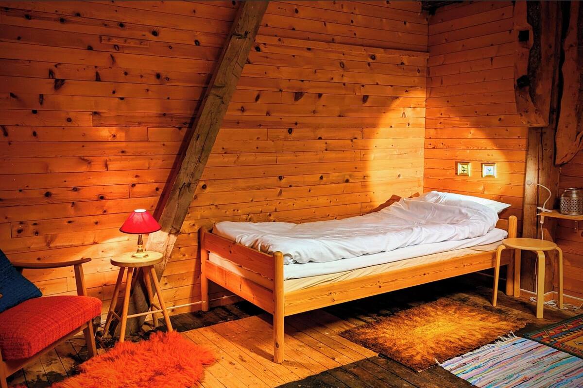 A cozy wooden cabin room with a single bed, a small table with a lamp, a chair, and a rug.