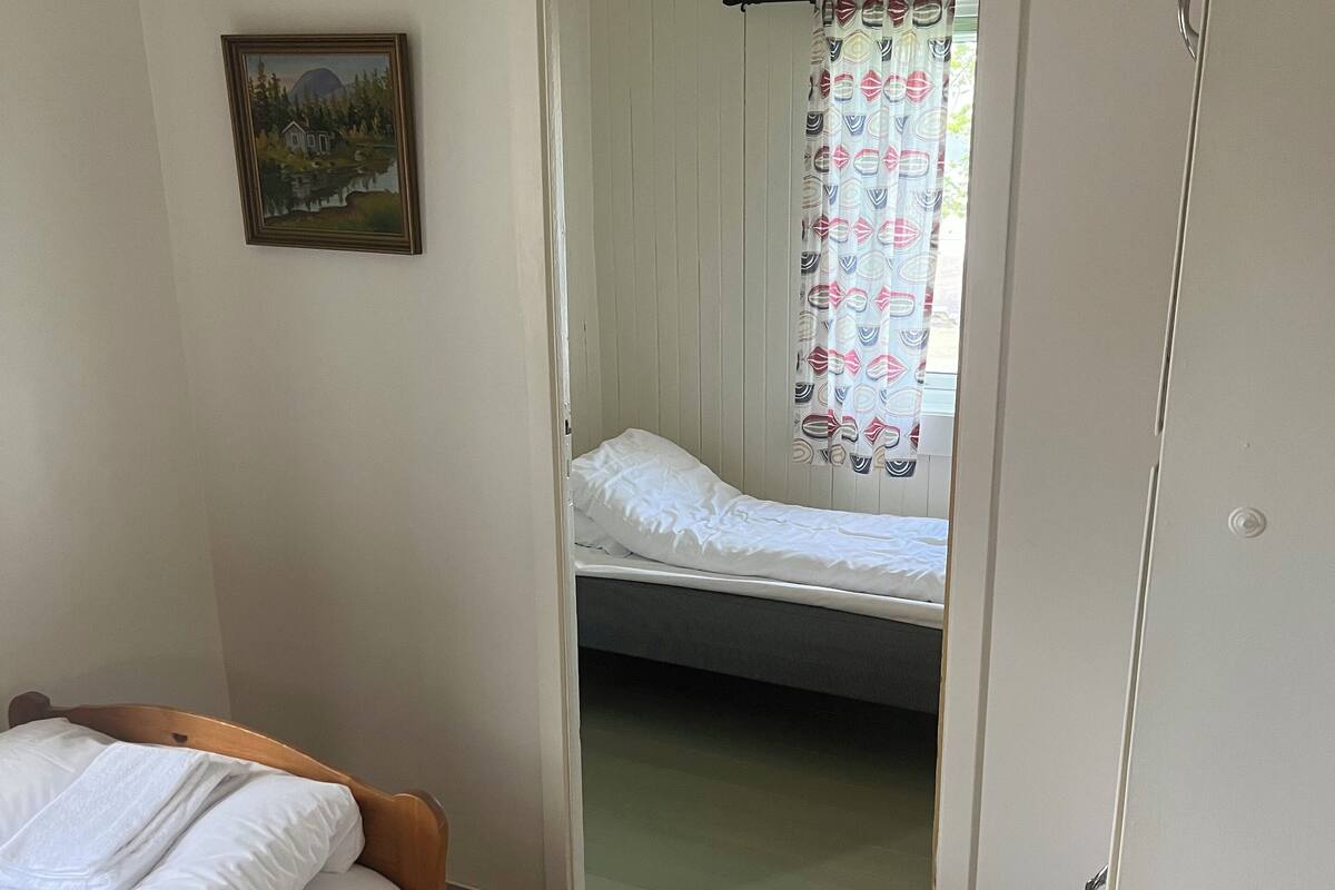 A bedroom with a wooden bed, a small bedside table, a framed picture on the wall, and a door leading to another room.
