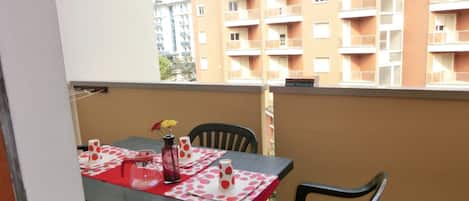 A balcony with a table set for two, featuring a red tablecloth and a vase with flowers.