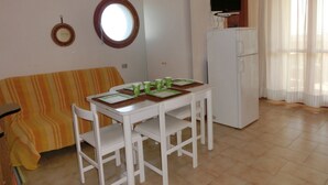 Ein Essbereich mit Tisch, Stühlen, einer Couch, einem Kühlschrank, einem Fernseher und einem Fenster mit Vorhängen.