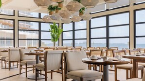 A spacious dining area with round tables and chairs, large windows offering a view, and hanging wicker light fixtures. - AVA Resort Cancun (Cancun)