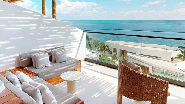 A balcony with a sofa, a wicker chair, and a small table. The balcony overlooks a body of water and a distant shoreline. - AVA Resort Cancun (Cancun)