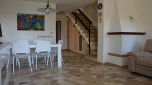 A dining area with a white table and chairs, a painting on the wall, a staircase, and a beige sofa.
