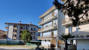 A multi-story residential building with balconies, a gravel driveway, and a fenced area with trees.
