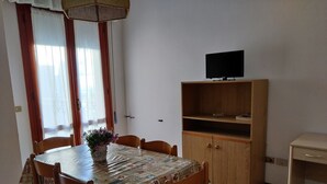 A dining area with a table covered by a patterned tablecloth, surrounded by wooden chairs. A television is mounted on a wooden cabinet above the table.