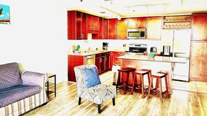 A compact kitchen with wooden cabinets, a microwave, a refrigerator, and a coffee maker. There is a bar stool and a patterned armchair in the living area. - PCB SNOWBIRDS PARADISE/GOLF/5POOLS/SOME HEATED/STEPS TO BEACH (Panama City Beach)