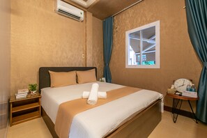 Una cama bien hecha con cubrecama blanco y beige, una mesita de noche con una lámpara, un reloj y un cartel que indica el tipo de habitación.