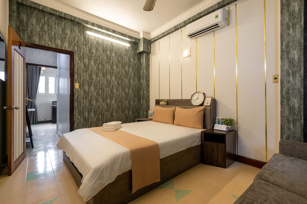 A hotel room with a large bed, a bedside table with a clock, a small plant, and a wall-mounted air conditioner.
