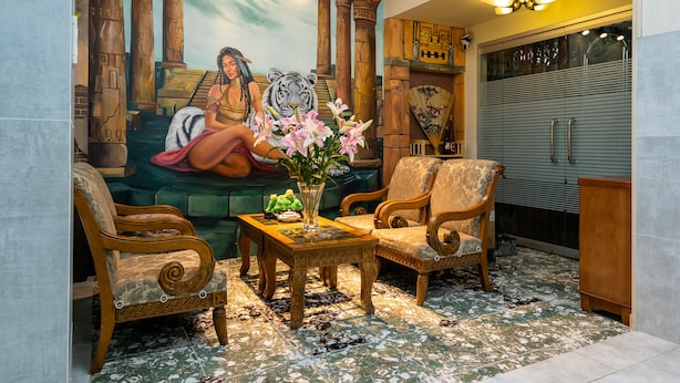 Quarto com um mural de uma mulher e um tigre, duas poltronas de madeira, uma mesa pequena com um vaso de flores e uma janela grande com persianas.