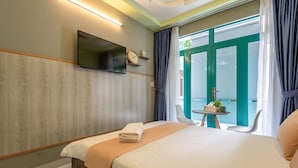 Quarto de hotel com cama, televisão presa na parede, pequena mesa com planta e vista para o exterior por uma porta de vidro.