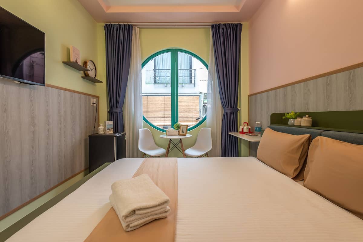 A hotel room with a bed, a desk, two chairs, a TV, and a window with curtains.