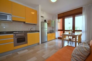 Une cuisine compacte avec des meubles de couleur jaune, un four à micro-ondes, une cuisinière et un réfrigérateur. On y trouve un coin repas avec une table et des chaises, ainsi qu’une fenêtre avec des rideaux.