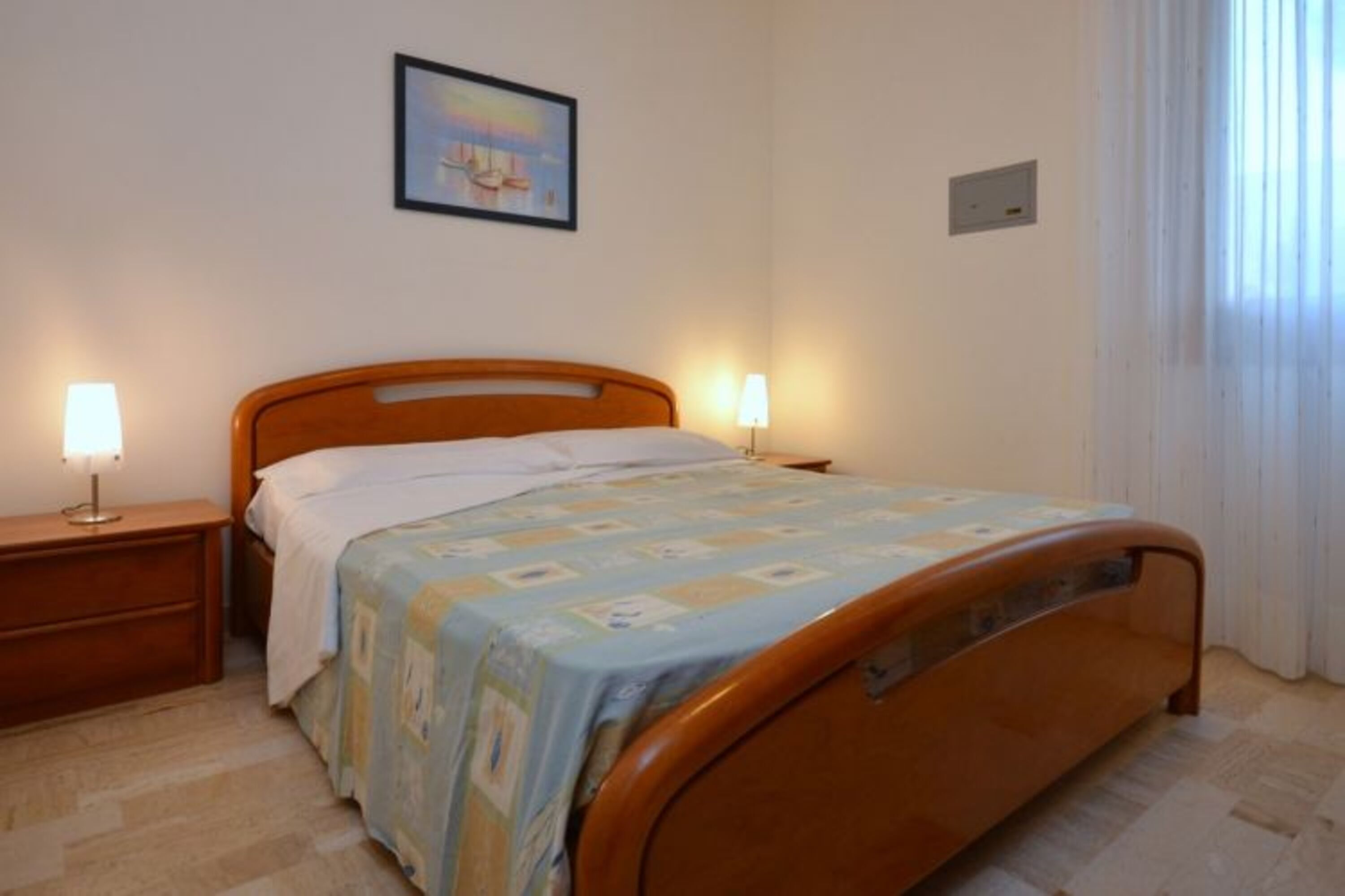 A bedroom with a wooden bed, two bedside tables with lamps, a framed picture on the wall, and a window with curtains.