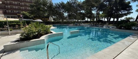 Una piscina con solárium empedrado, rodeada de árboles y un edificio al fondo.