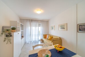 A compact living space with a dining table, a sofa, a kitchenette, and a window with curtains. - Colourful and lively apartment in Lignano (Lignano Sabbiadoro)
