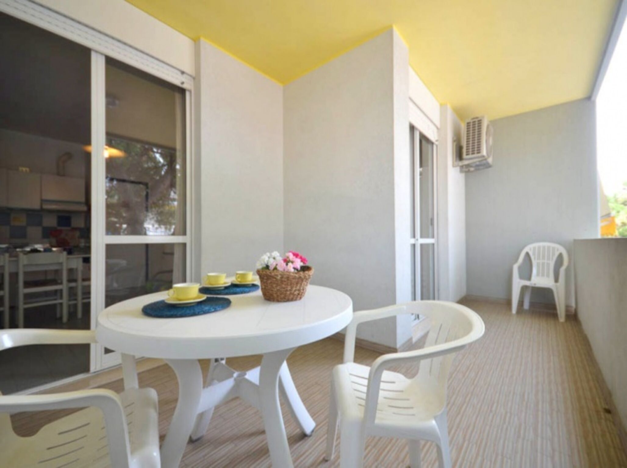 A small outdoor dining area with a white table and chairs, a basket of flowers, and a view of the kitchen through a glass door.