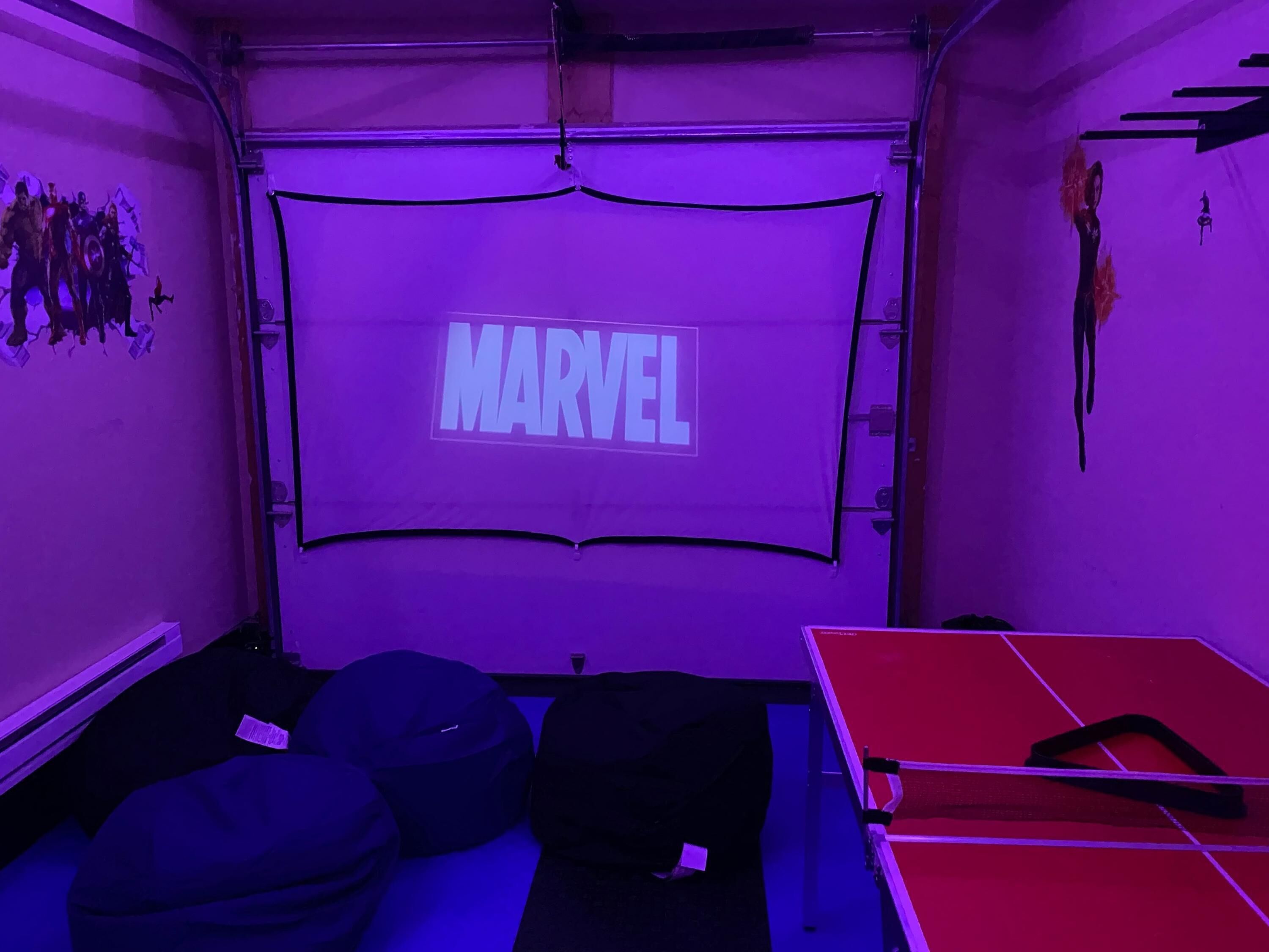 Een kamer met een groot scherm waarop 'MARVEL' staat, zitzakken, een pingpongtafel en een muur met posters van superhelden.