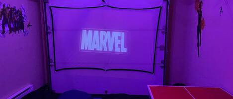 Een kamer met een groot scherm waarop 'MARVEL' staat, zitzakken, een pingpongtafel en een muur met posters van superhelden.