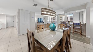 A modern kitchen with a central island, a dining area with a glass table, and a living space with a sofa and TV. - Gullwing Resort 1002- A beachfront condo with amazing beach and bay views (Fort MyersBeach)