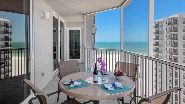 A balcony with a table set for two, overlooking a beach and high-rise buildings. - Gullwing Resort 1002- A beachfront condo with amazing beach and bay views (Fort MyersBeach)