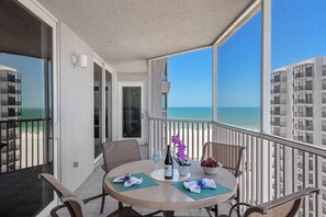A balcony with a table set for two, overlooking a beach and high-rise buildings. - Gullwing Resort 1002- A beachfront condo with amazing beach and bay views (Fort MyersBeach)