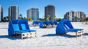 A beach with sun loungers covered in blue tarps, palm trees, and high-rise buildings in the background. - Gullwing Resort 1002- A beachfront condo with amazing beach and bay views (Fort MyersBeach)