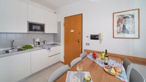 Una cocina moderna con armarios blancos, un microondas y una cafetera. Hay una mesa de comedor con una botella de vino, una copa y un poco de pan. Un cuadro enmarcado cuelga en la pared.