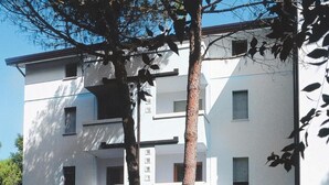 A multi-story residential building with a flat facade, balconies, and a small front yard with a lawn and trees. - Comfortable apartment in a quiet area (Bibione Pineda)