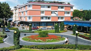 Um hotel de vários andares com fachada vermelha e branca, um pátio central com flores vermelhas e um toldo azul com o nome 'Gelateria'.