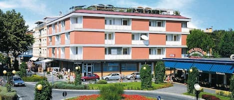 Un hôtel à plusieurs étages, doté d’une façade rouge et blanche, d’une cour intérieure ornée de fleurs rouges et d’un auvent bleu sur lequel est inscrit le nom « Gelateria ».