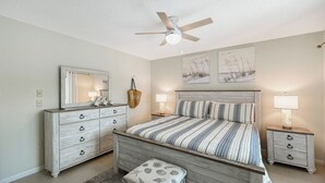 Une chambre à coucher comprenant un lit, des tables de chevet, une commode, un ventilateur de plafond et des œuvres d’art murales.