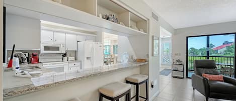 Your open-concept kitchen flows seamlessly into the living area, with granite counters, bar seating, and views that stretch out to a sunny balcony and tree-lined horizon beyond.