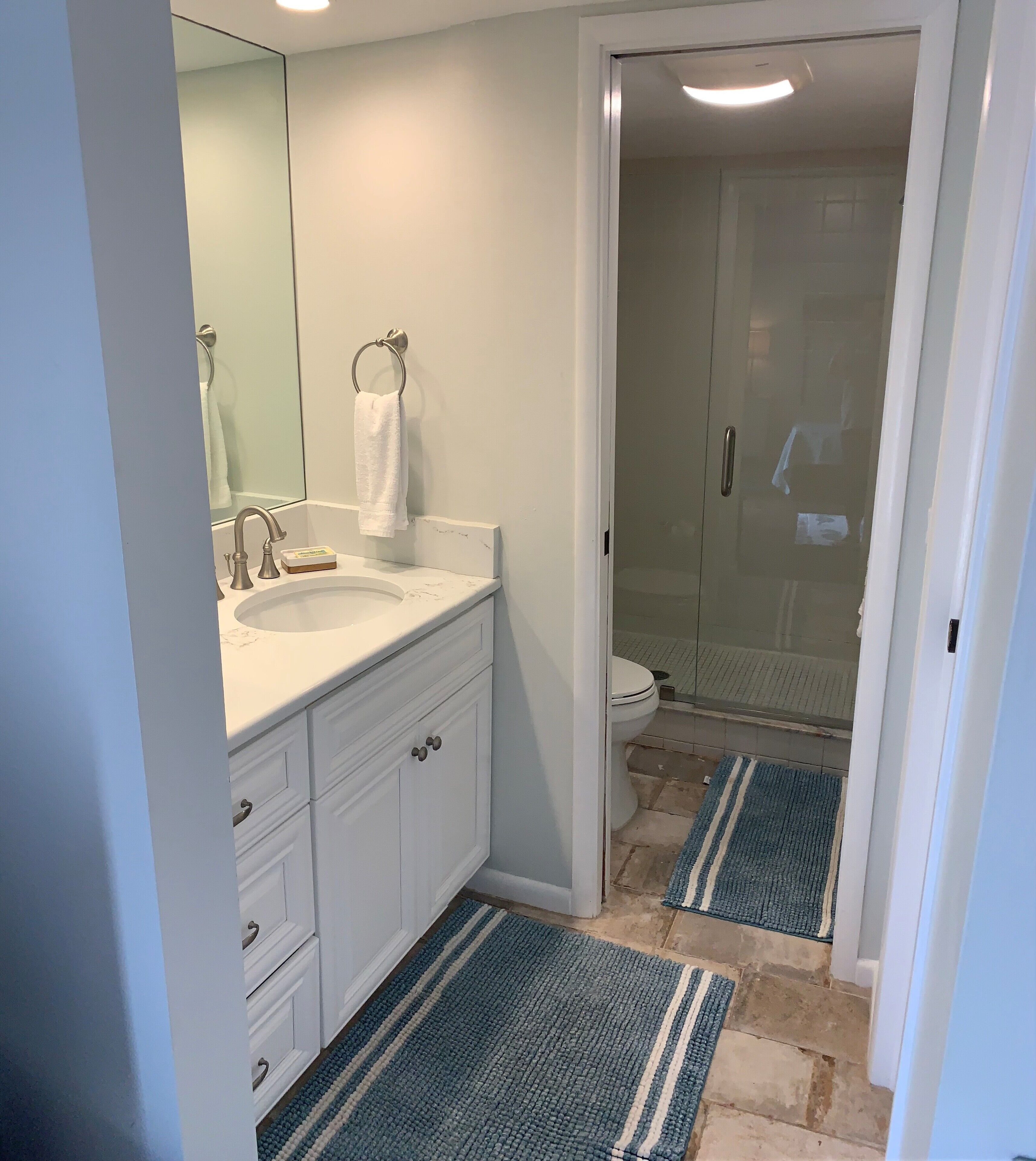 A bathroom with a white sink, mirror, and cabinet. There is a towel hanging on a ring, and a shower area with a glass door. Blue and white striped rugs are on the floor.