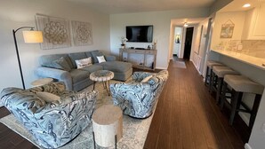 A modern living room with a grey sectional sofa, patterned ottoman, and a small coffee table. A flat-screen TV is mounted on the wall, and there is a kitchen area with bar stools visible. - Spectacular view on Crescent beach (Sarasota)