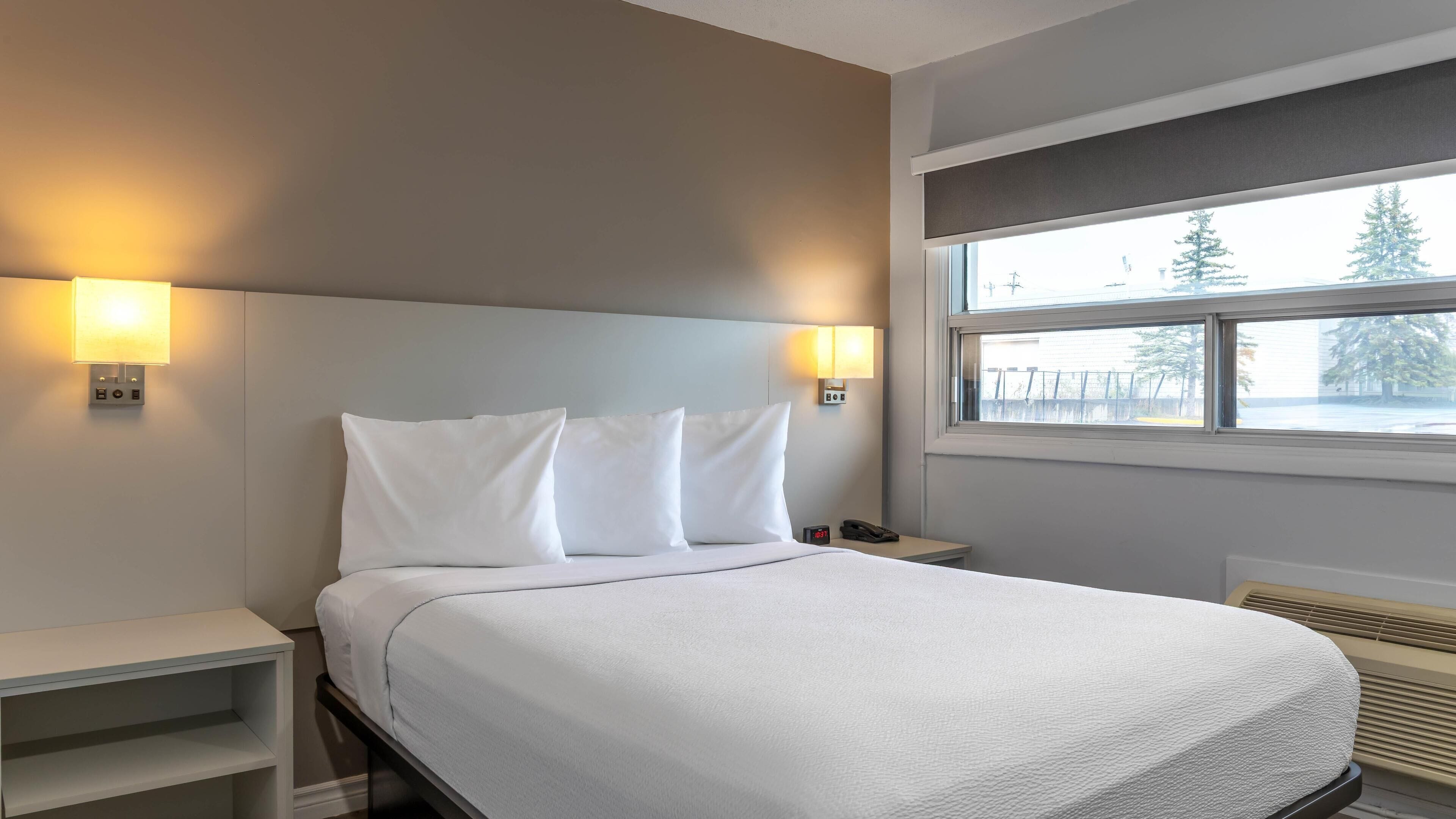 Habitación de hotel con cama, mesitas de noche, lámparas fijadas a la pared, ventana con vistas a árboles y termostato.