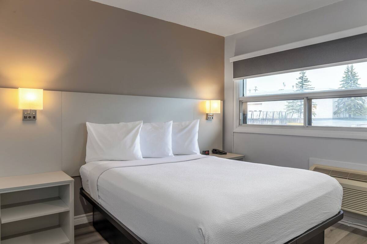 A hotel room with a bed, bedside tables, a nightstand, a window with a view of trees, and wall-mounted lights.
