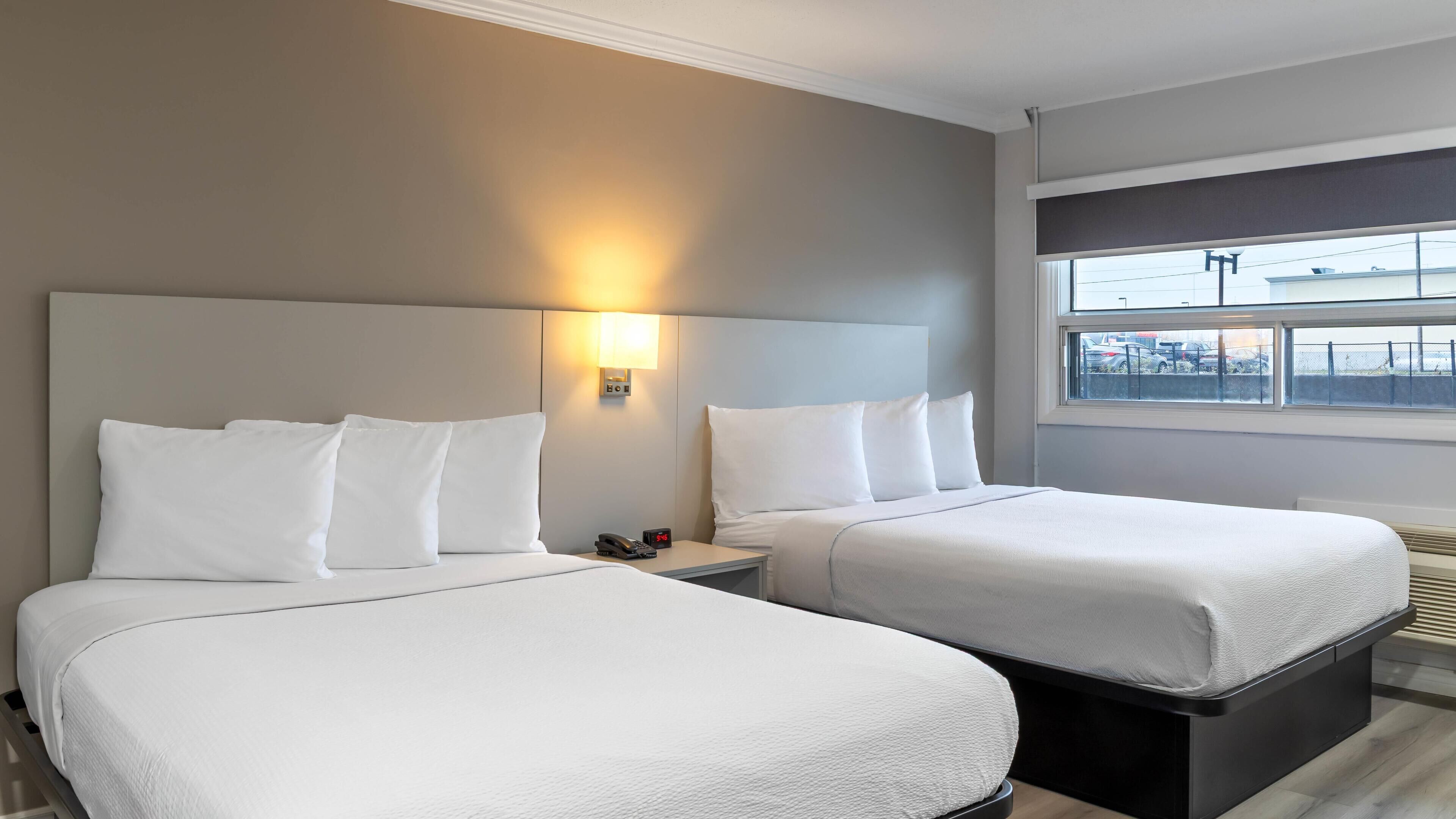 Une chambre d’hôtel avec deux lits, une table de chevet avec un téléphone et un réveil, une lampe fixée au mur et une fenêtre donnant sur l’extérieur.