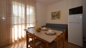 A small, well-lit room with a dining table set for two, a sofa, a refrigerator, and a painting on the wall.