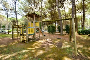 Un espace de jeux avec une structure d’escalade en bois et des balançoires, situé dans un parc arboré.
