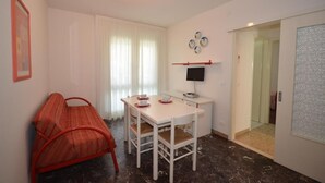 Ein kleiner, gut beleuchteter Raum mit einem weißen Tisch und Stühlen, einem roten Sofa, einem Fernseher auf einem Schrank und einer Tür zu einem weiteren Zimmer.